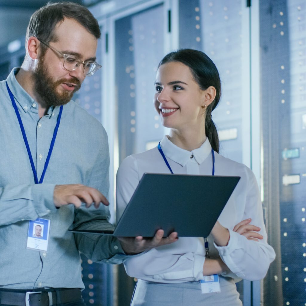 Bearded IT Technician in Glasses and Beautiful Young Engineer Colleague with a Laptop Computer are Talking in Data Center while Working Next to Server Racks. Running Diagnostics or Doing Maintenance Work.
